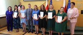 Wojewoda Opolski, Opolski Kurator Oświaty oraz Opolski Wicekurator Oświaty stoją z częścią awansowanych nauczycieli. Wszyscy pozują do zdjęcia grupowego. Nauczyciele mają w rękach otrzymane akty nadania stopnia awansu. Ściany w tle są żółte, stoją tam również flagi Polski oraz Unii Europejskiej. 