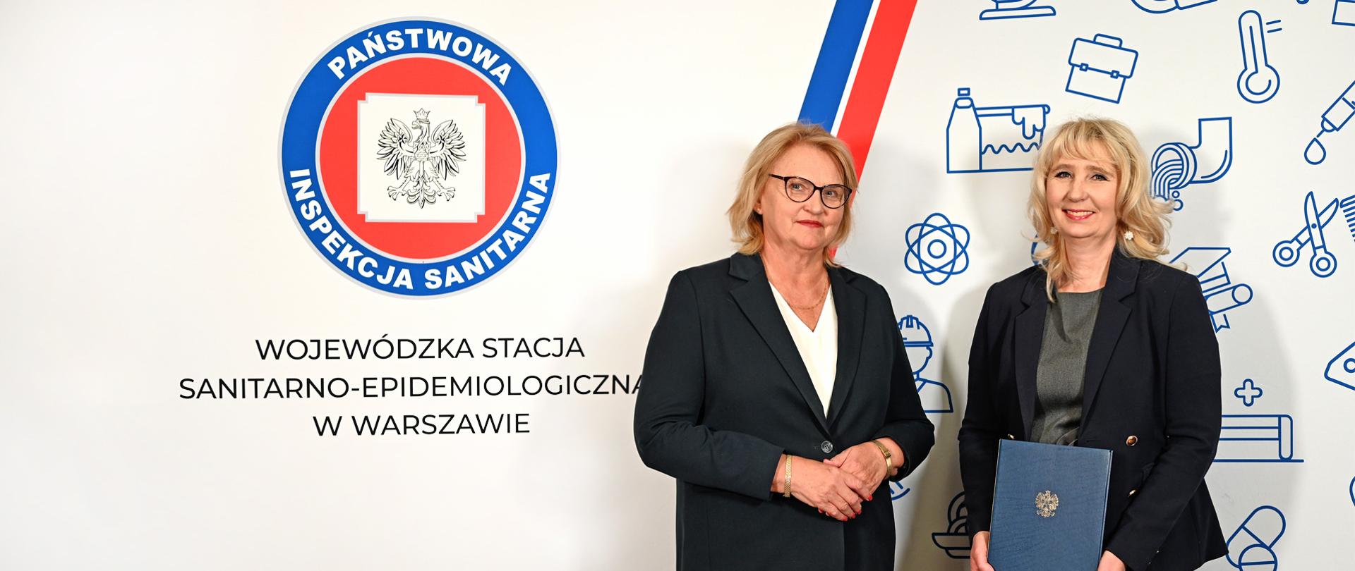 MPWIS Elżbieta Stefaniuk wręcza akt powołania dla Zastępcy MPWIS Marzeny Woźniak na tle ścianki z logo Państwowej Inspekcji Sanitarnej.