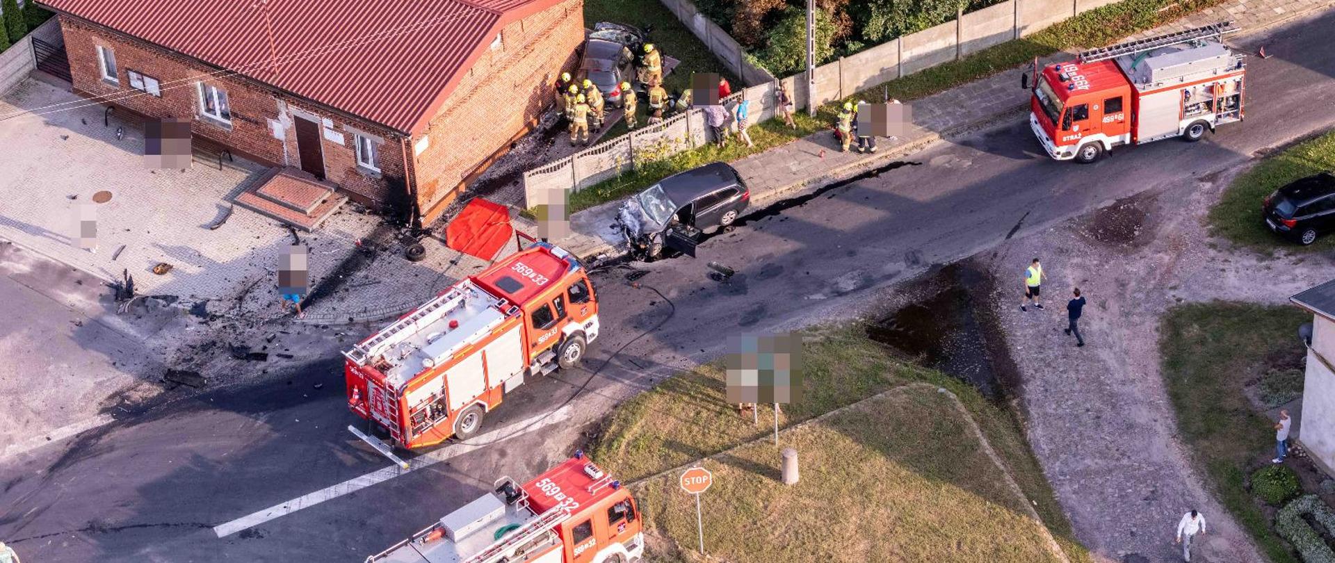 Zdjęcie wykonane z drona przedstawia służby ratunkowe realizujące działania na skrzyżownaiu dróg gdzie doszło do zderzenia dwóch samochodów osobowych. Jeden z uszkodzonych pojazdów widoczny na poboczy drogi, drugi przy budynku na jednej z posesji. Strażacy realizują działania. Droga całkowicie zablokowana. Widoczne trzy zastępy pożąrnicze blokujące calkowicie przepustowość drogi