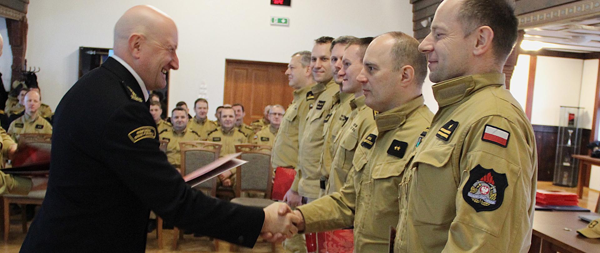 Sześciu strażaków w piaskowych mundurach służbowych, stoją w szeregu. Podszedł do nich strażak w mundurze wyjściowym i przekazuje dokument jednemu z nich. W tle siedzą inni, na krzesłach, w rzędach... Widać ozdobne elementy konstrukcyjne sali.