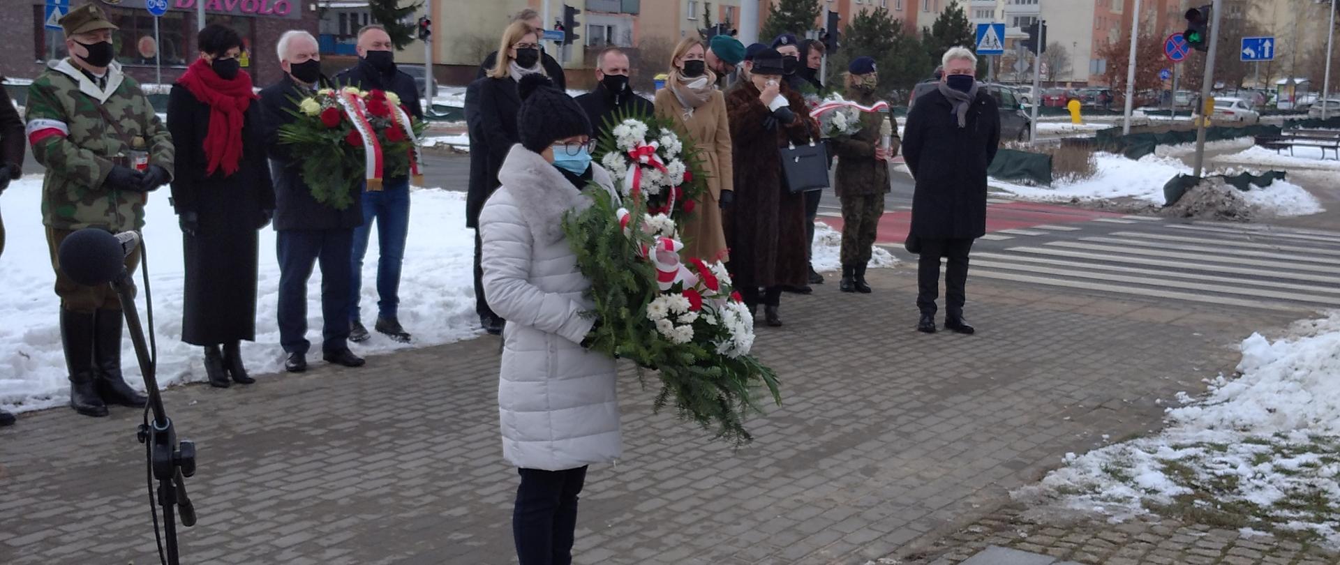 Na pierwszym planie znajduje się kobieta trzymająca w ręku wieniec. W tle w szeregu stoi grupa ludzi, dwoje z nich również trzyma wieńce