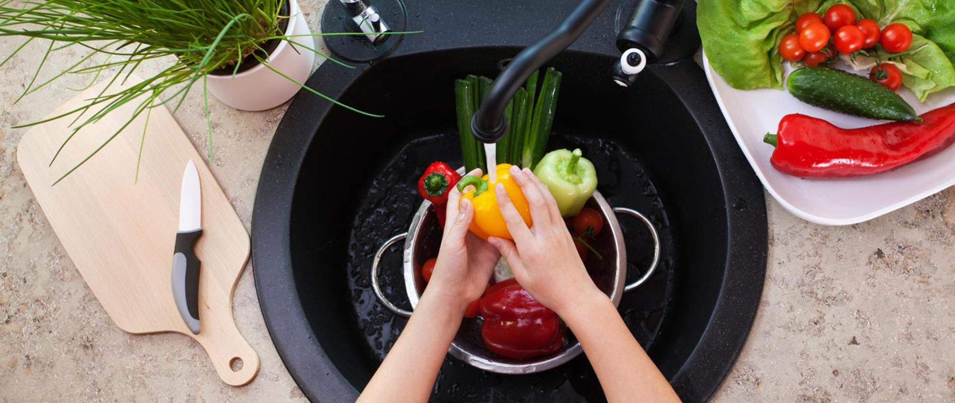 Po lewej deska do krojenia, na niej nóż, powyżej doniczka ze szczypiorkiem, na środku czarny zlew i kran, rece myją kolorowe papryki, po prawej na talerzu zielona sałata, pomidorki, ogórek, czerwona papryka