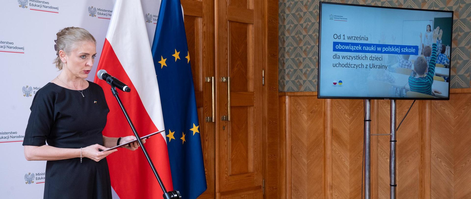 Na tle białej ścianki z napisami Ministerstwo Edukacji Narodowej oraz flag Polski i UE stoi przed ustawionym na statywie mikrofonie wiceminister Joanna Mucha. W ręku trzyma bordową teczkę. Po prawej stronie na statywie ustawiony monitor na którym wyświetlana jest prezentacja. 