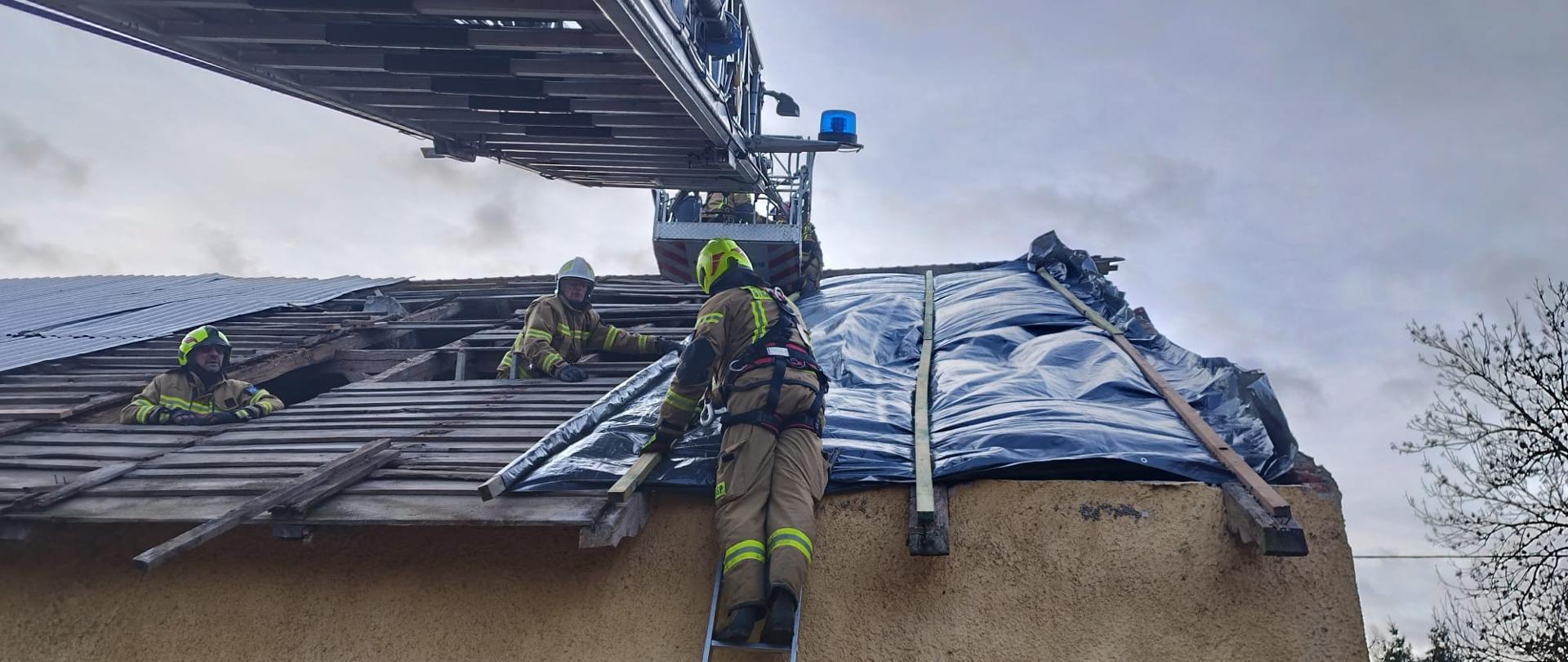 Uszkodzony dach. Na dachu strażacy nakładają plandekę. U góry zdjęcia drabina mechaniczna.