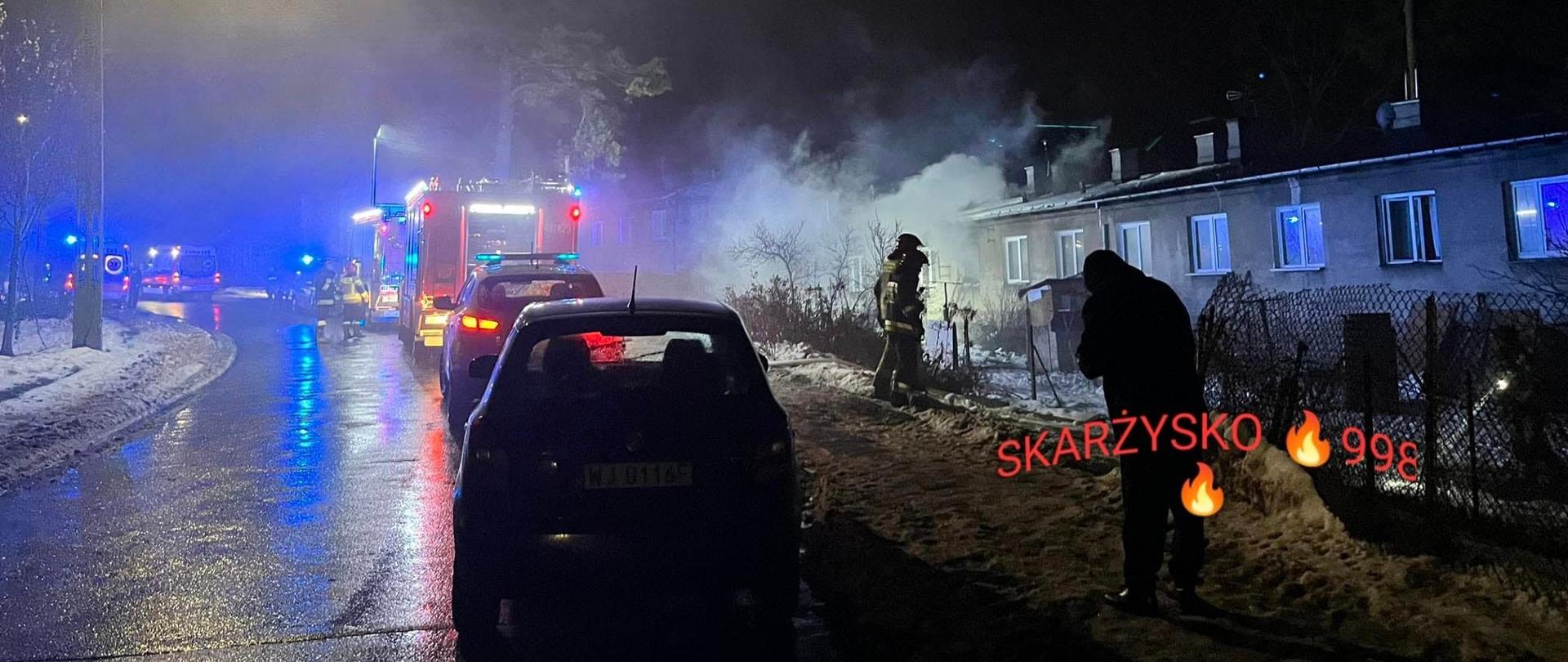 Widok na akcję gaśniczą podczas pożaru budynku mieszkalnego. Z budynku wydobywa się zadymienie, na drodze stoją samochody strażackie.