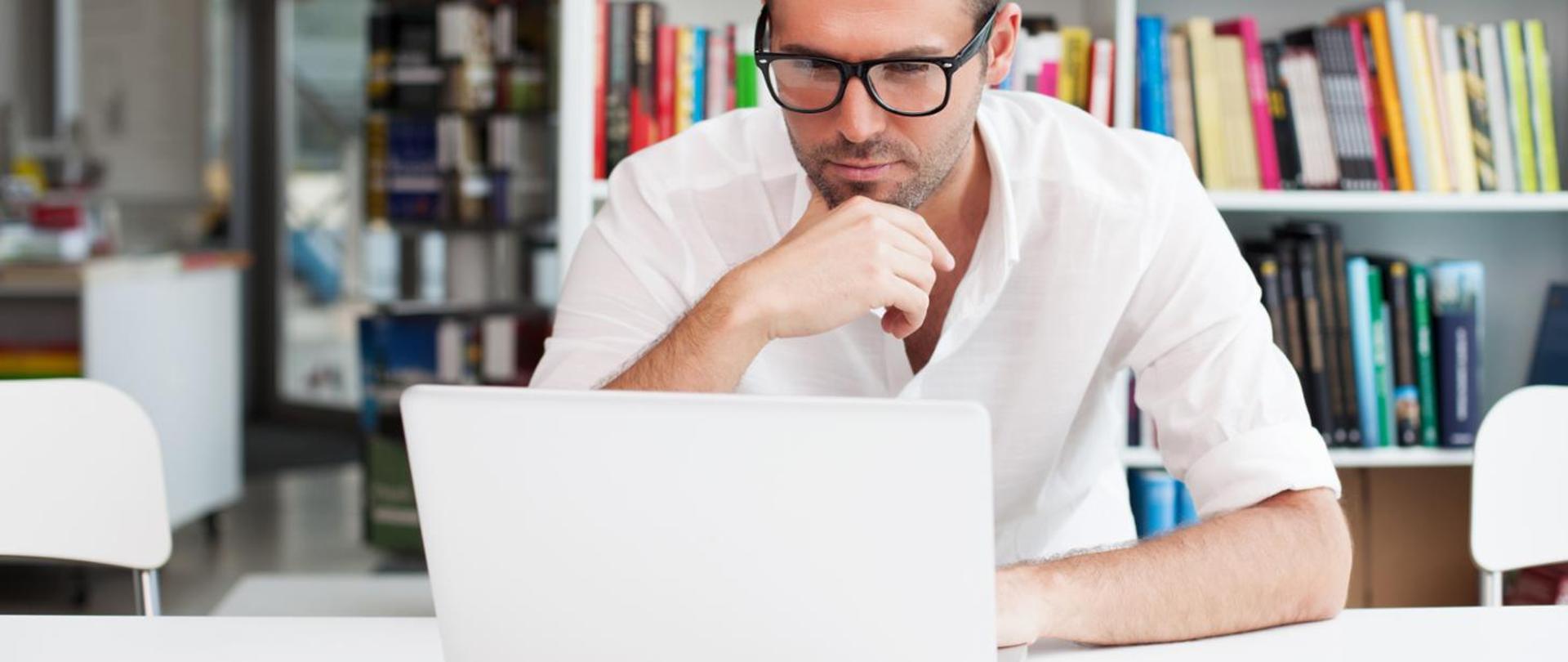 Zamyślony mężczyzna w okularach, ubrany na biało, siedzi przy stole i wpatruje się w ekran laptopa. Za nim regał z kolorowymi książkami oraz białe biurko.
