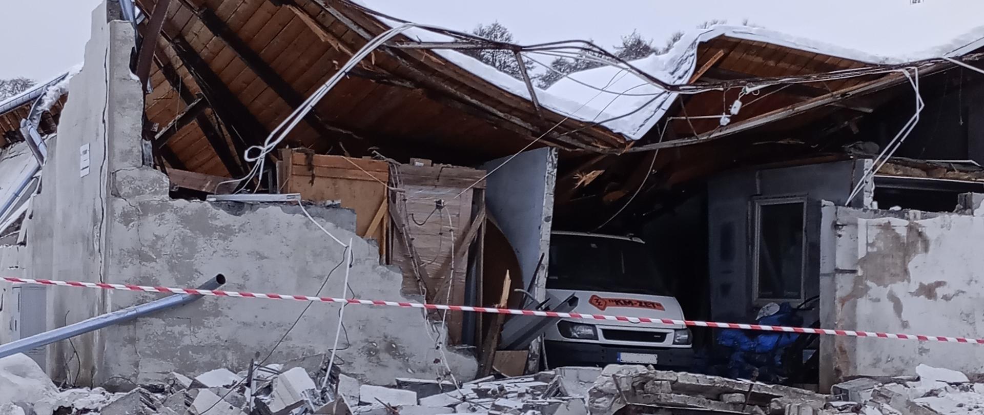 Na pierwszym planie widoczny jest budynek z zawalonymi ścianami oraz dachem na którym widać pokrywę śnieżną. Przed budynkiem widoczny jest śnieg natomiast w jego wnętrzu stoi biały samochód typu mikrobus. Budynek ogrodzony jest dookoła biało-czerwoną taśmą. Zdjęcie powiększy się po kliknięciu na nie.