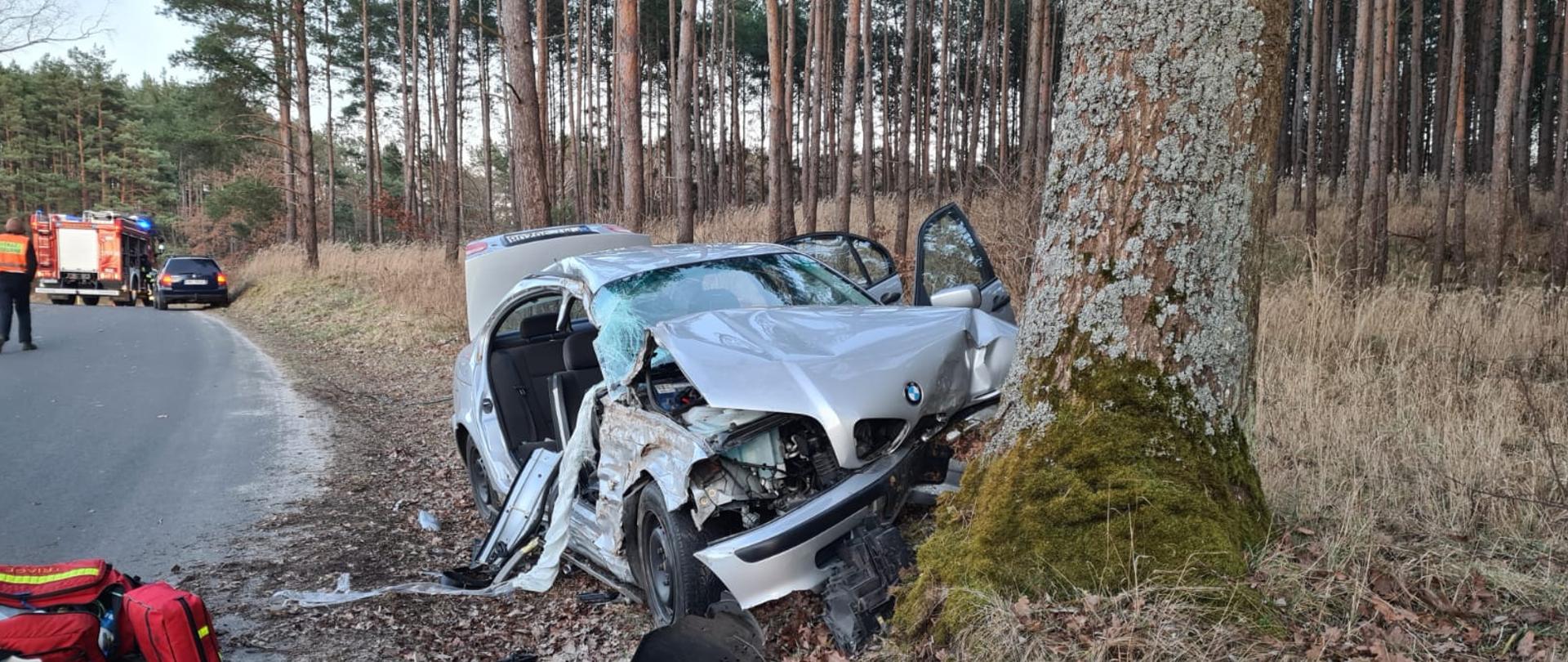 Zdjęcie przedstawia rozbity na drzewie pojazd osobowy marki BMW. Obok pojazdu znajduje się strażacki zestaw do pomocy medycznej R-1 PSP. W tle pojazd strażacki średni gaśniczy z modułem technicznym.