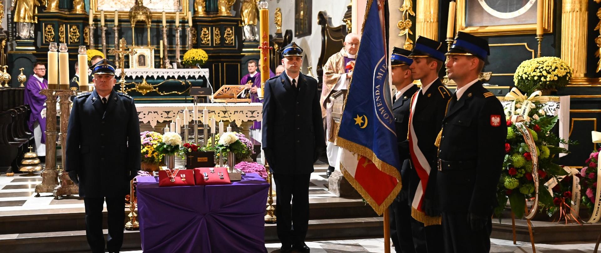 Obok urny po prawej i po lewej stronie stoją strażacy ubrani w mundury galowe, urna stoi na fioletowym postumencie, obok urny kwiaty oraz odznaczenia. Z tyłu ołtarz oraz świece. Po prawej stronie stoi poczet sztandarowy trzech strażaków branych w granatowe mundury galowe. Środkowy strażak trzyma sztandar.