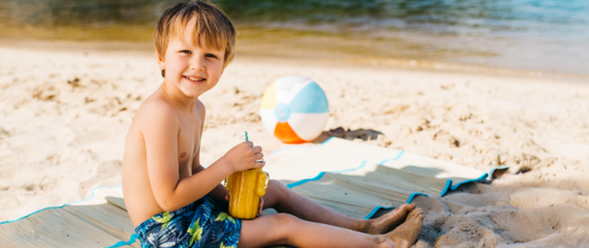 Dziecko, chłopczyk siedzi na plaży trzymając kubek . W tle piłka plażowa oraz brzeg akwenu