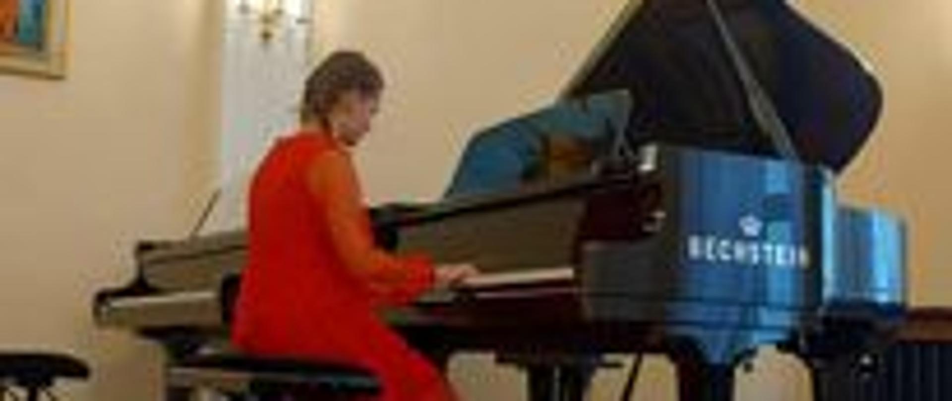 Zdjęcie przedstawia kobietę w czerwonej sukni, grającą na fortepianie na sali koncertowej