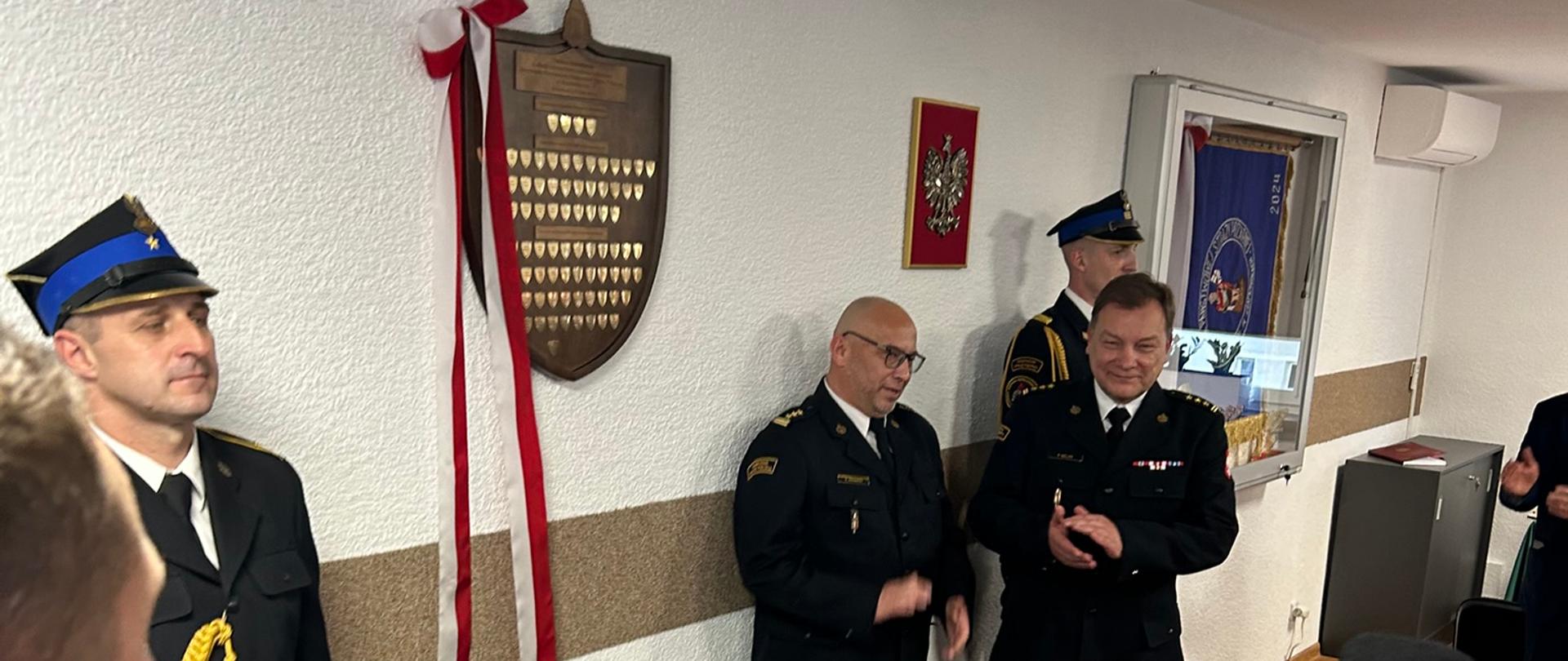 3. Na świetlicy KP PSP w Krapkowicach na ścianie zawieszona pamiątkowa tablica z gwoździami symbolizującymi darczyńców. Pod tablica na stole okrytym zielonym suknem biało-czerwona wiązanka kwiatów. Po bokach tablicy dwóch strażaków w umundurowaniu galowym, pełni posterunek honorowy . Obok tablicy stoją Opolski Komendant Wojewódzki PSP i Komendant Powiatowy PSP w Krapkowicach, którzy przed chwilą dokonali odsłonięcia tablicy . 