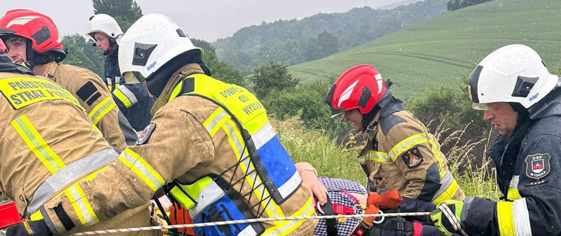 Na zdjęciu znajdują się strażacy wchodzących przy pomocy lin po skarpie, którzy transportują poszkodowanego na noszach. W tle widać pola