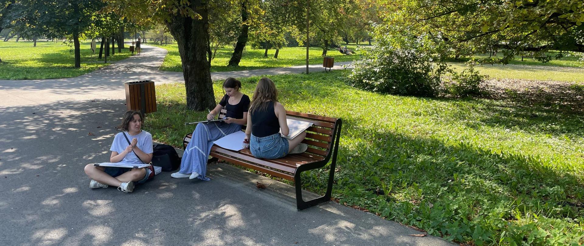 Jest piękna, słoneczna pogoda. Troje uczniów w Parku Ludowym w Lublinie. Wykonują rysunki. Dwie uczennice siedzą na ławce. Jedna przodem w pozycji normalnej. Druga siedzi tyłem do kadru. Ma skrzyżowane nogi. Trzecia uczennica siedzi na asfalcie. Ma skrzyżowane nogi. W tle roślinność, drzewa, trawniki oraz asfaltowe alejki.