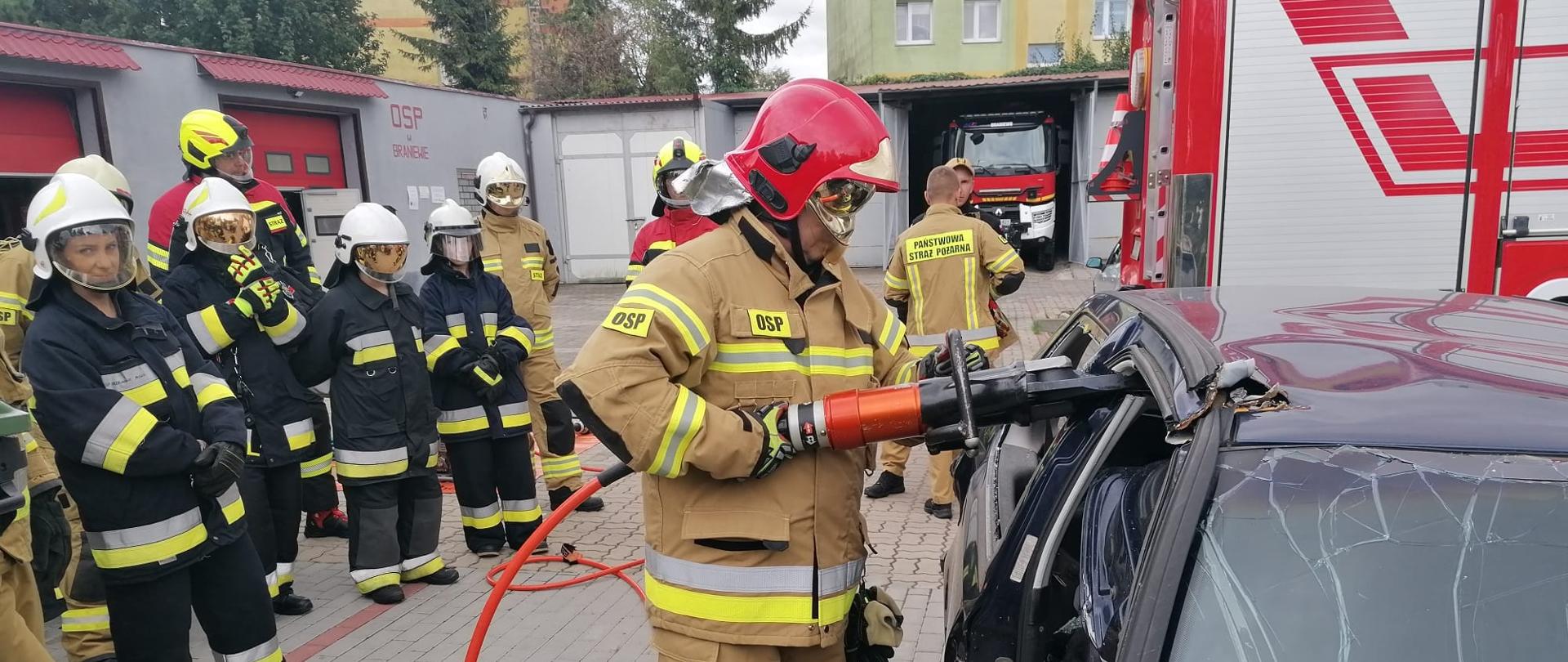 Grupa strażaków w mundurach bojowych i hełmach, trwa szkolenie.