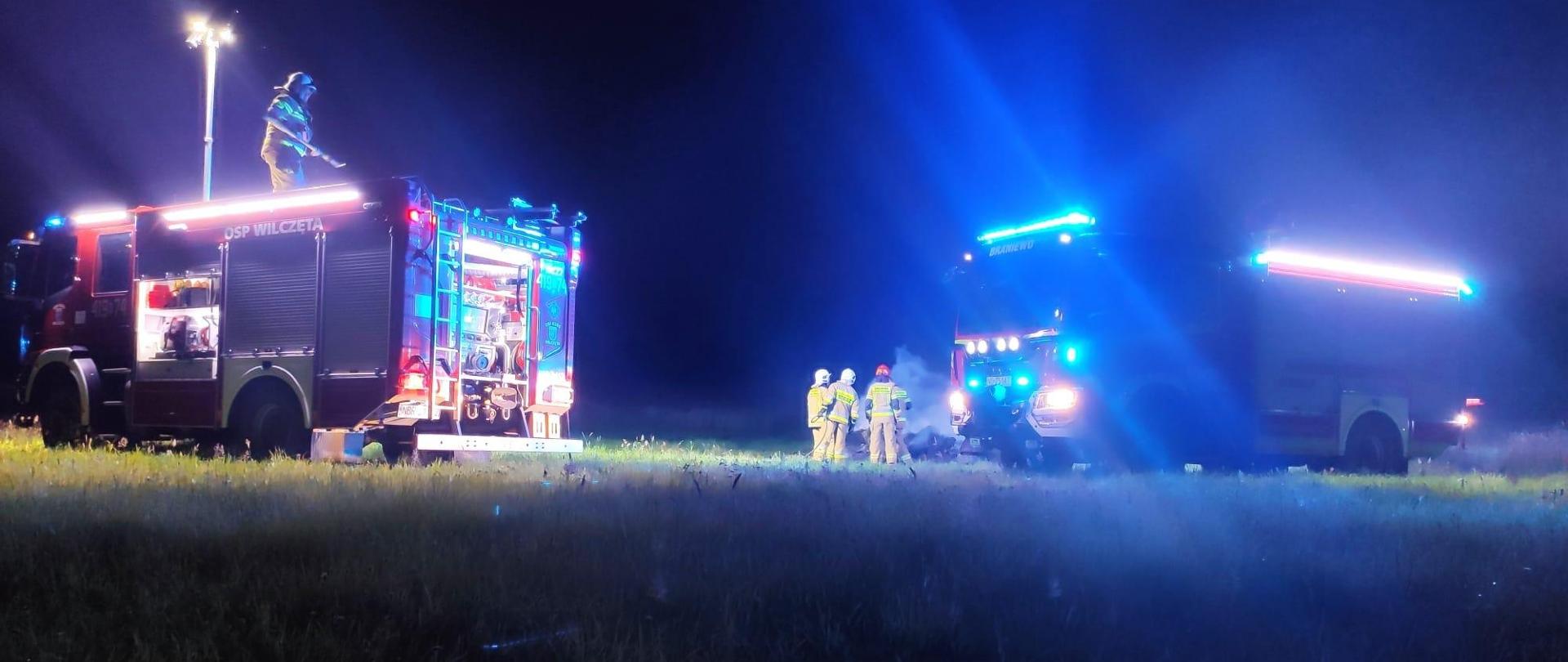 Zdjęcie zrobione nocą. Na zdjęciu dwa samochody strażackie z włączonymi światłami ostrzegawczymi niebieskimi. Przy samochodach widać strażaków.