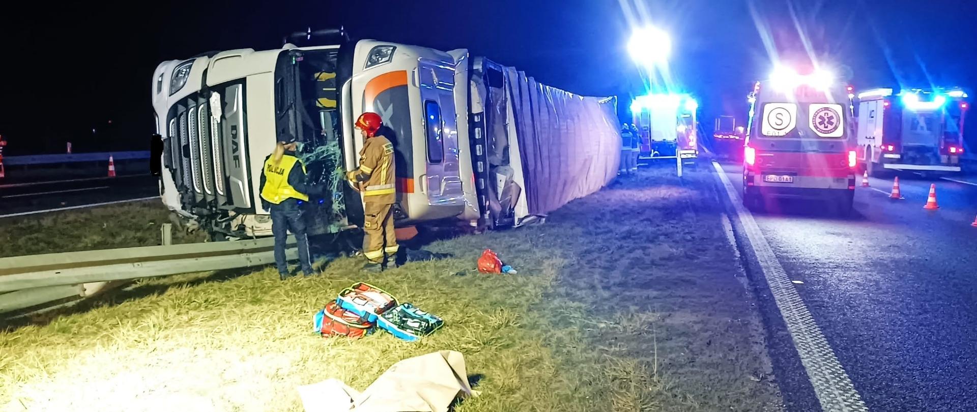 duzy samochód ciężarowy leży obok drogi przewrócony na bok, przednia szyba jest popękana, stoją przy niej strażak i policjantka 