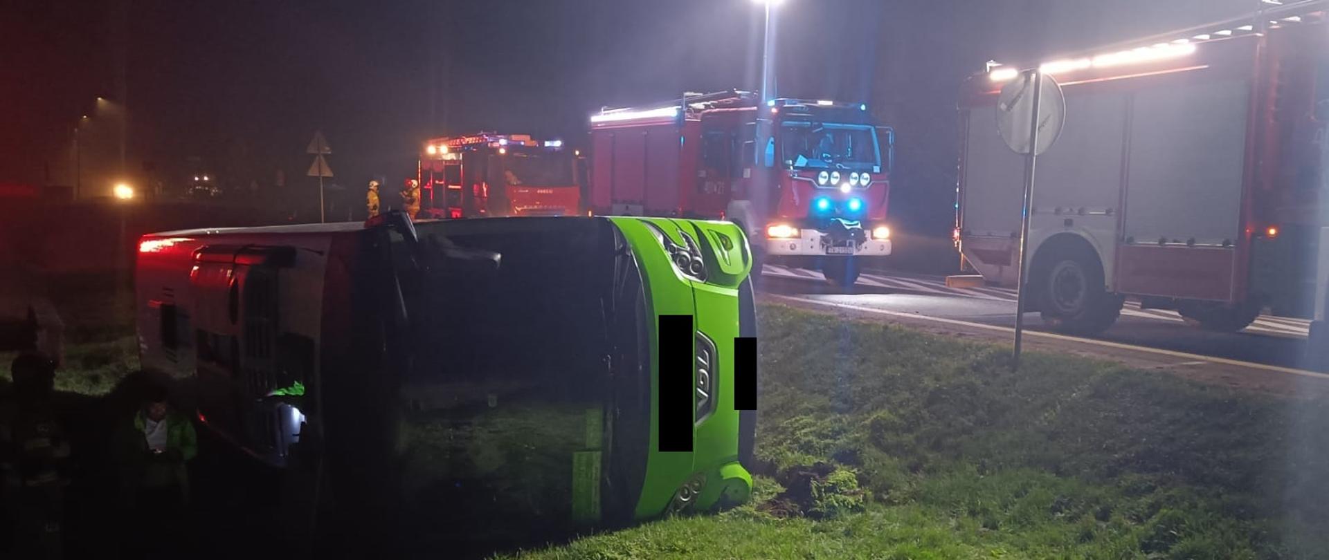 Po lewej stronie przewrócony zielony autokar, po prawej stronie samochody pożarnicze. Z tyłu po prawej stronie stoją strażacy PSP i OSP. 