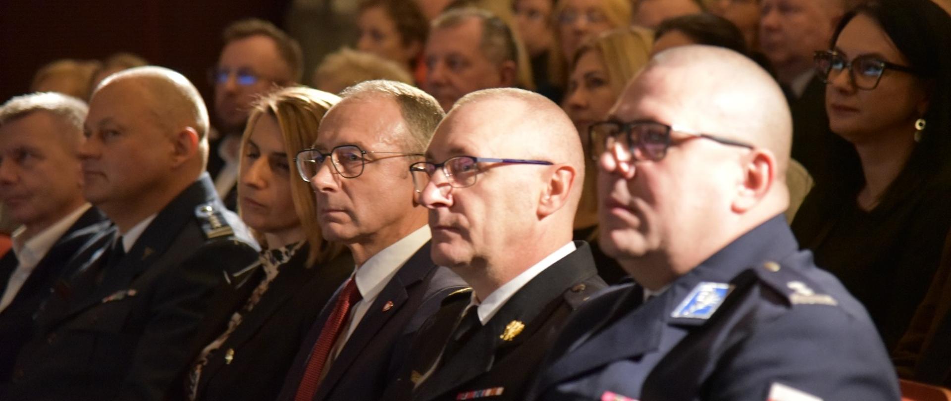 Na pierwszym planie lubuski komendant wojewodzki PSP obok wojewody lubuskiego i komendanta wojewodzkiego Policji. Siedzą w filharmonii. W tle inne osoby zaproszone. Słabe światło.