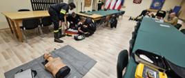 Na zdjęciu widoczna sala wykładowa wraz z uczestnikami egzaminu recertyfikacyjnego KPP, którzy przygotowują się do egzaminu praktycznego, na pierwszym planie widać po prawej ćwiczebny defibrylator AED, na środku widoczny fantom do ćwiczenia resuscytacji.