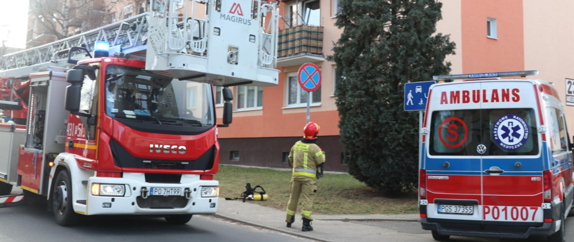 Strażaka stojącego pomiędzy drabiną mechaniczną a karetka pogotowia. Strażak zwrócony jest w kierunku bloku gdzie doszło do pożaru. Pojazdy mają włączone sygnały błyskowe.