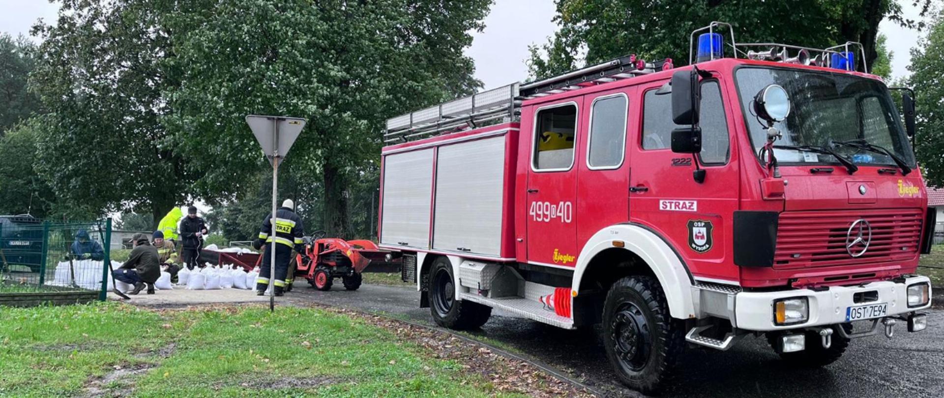 Na zdjęciu znajdują się strażacy w ubraniach specjalnych i osoby cywilne napełniające worki piaskiem, obok stoi samochód strażacki