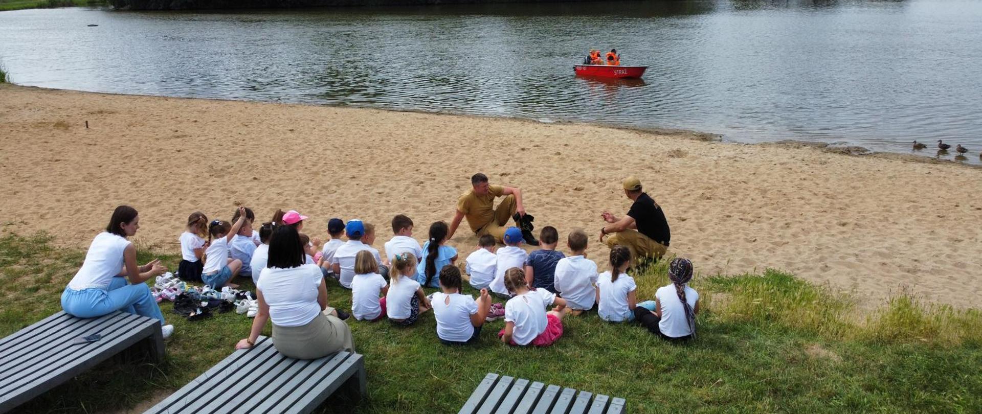 Grupa dzieci zasiada na plaży. Korzystając ze spotkania w tak szerokim gronie strażacy edukują najmłodszych przypominając podstawowe zasady bezpiecznego wypoczynku nad wodą. W tle łódź ratownicza ze strażakami, którzy doskonalą umiejętności manewrowania sprzętem