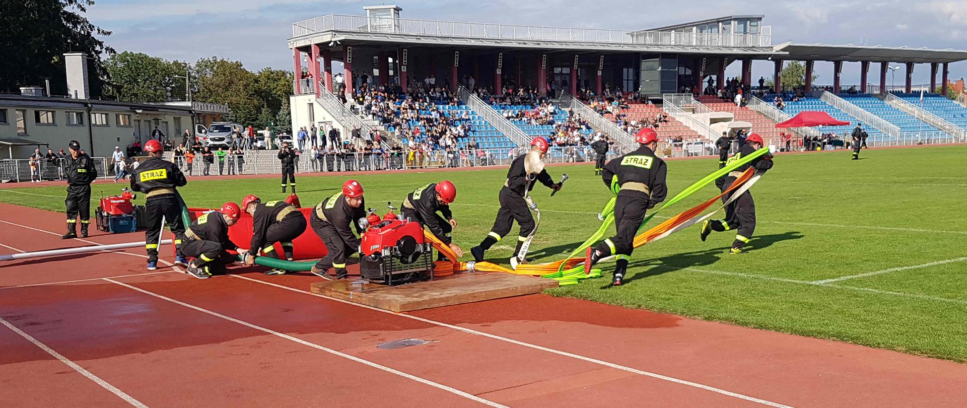 Zdjęcie zrobione w dzień na stadionie w Sandomierzu. Na zdjęciu widać strażaków - zawodników podczas łączenia sprzętu do konkurencji. Widać zbiornik z woda motopompę i w oddali wiadać inne drużyny. 