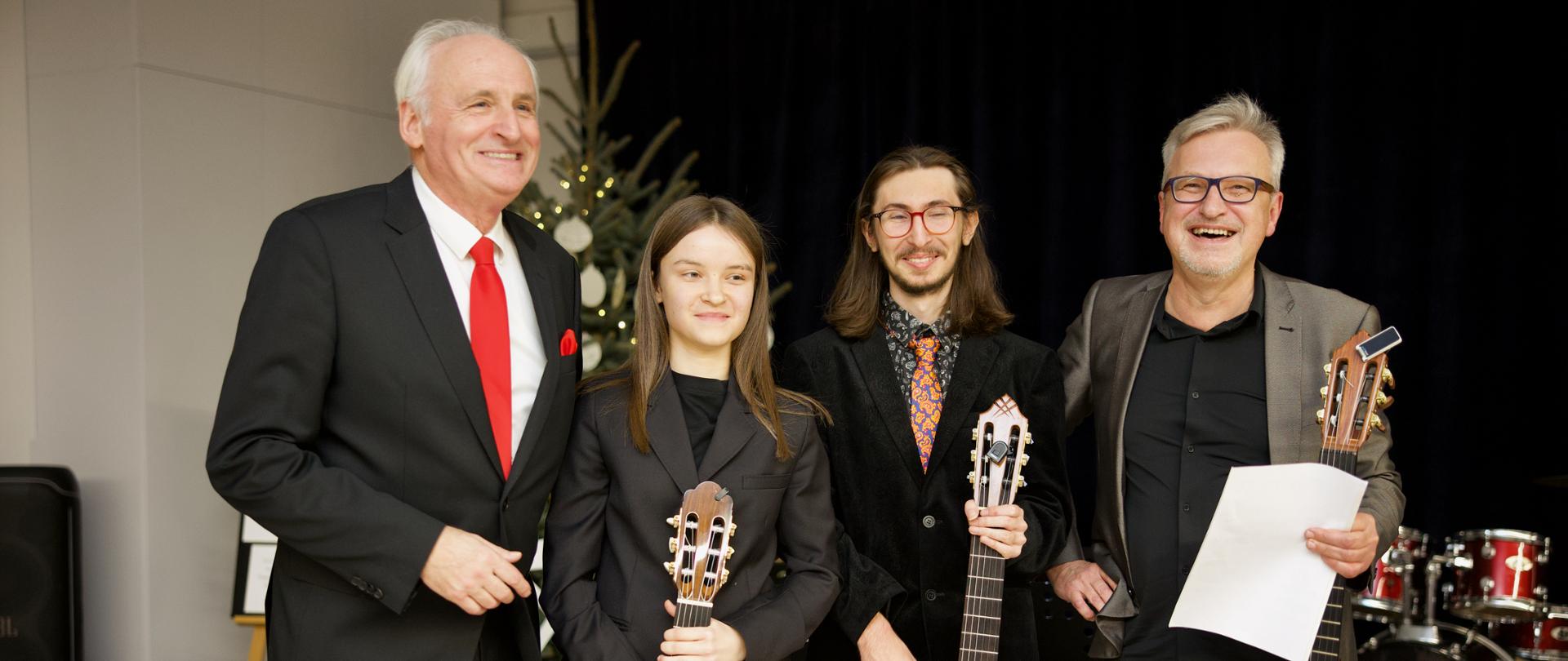 Zdjęcie z koncertu tria gitarowego. Trójka gitarzystów stoi, trzymając w rękach gitary. Od prawej: Andrzej Fil, Fryderyk Kłos, Gabriela Kościk. Po lewej, obok gitarzystów stoi Krzesimir Dębski. Cała czwórka uśmiechnięta i zadowolona. A. Fil trzyma w ręku nuty z autorskim opracowaniem utworu K. Dębskiego, wykonanego chwilę przed zrobieniem zdjęcia. W tle widać elementy sceny m. in. zestaw perkusyjny, krzesła i choinkę.