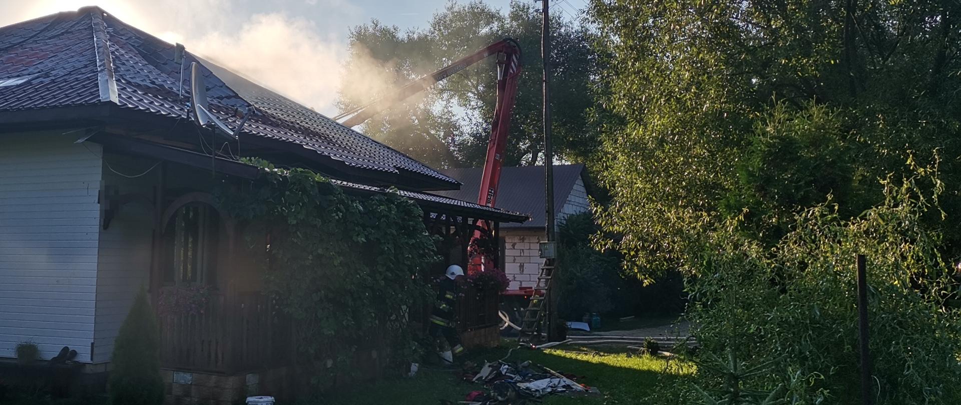 Na pierwszym planie widać palący się budynek. W tle widać strażaków, oraz rozłożony podnośnik straży pożarnej.