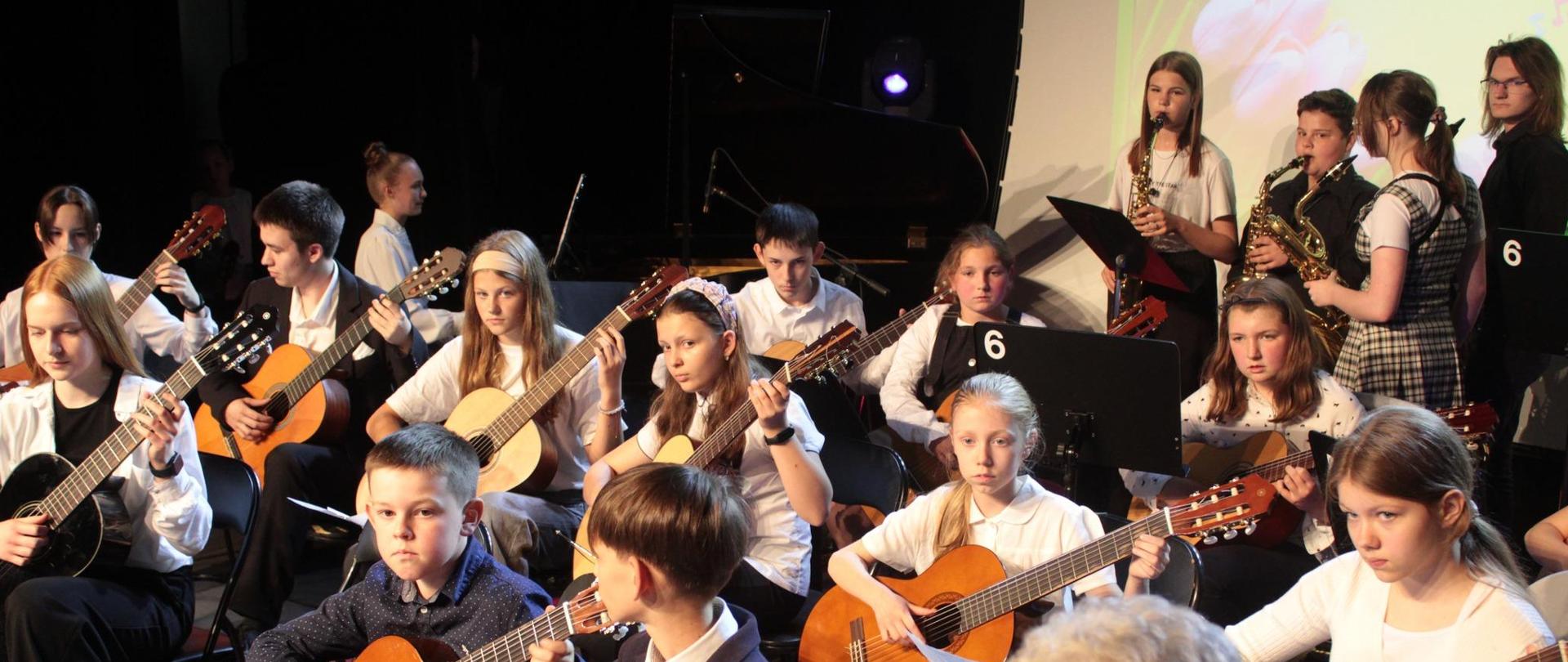 Uczniowie tworzący zespół siedzą, grając na gitarach. Z tyłu stoją uczniowie grający na saksofonach.
