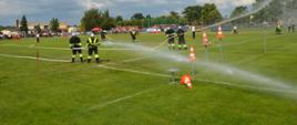 XI Powiatowe Zawody Sportowo-Pożarnicze OSP i MDP. Stadion w Miejskiej Górce. Jedna z drużyn jest na końowym etapie ćwiczenia bojowego. Roty przy pomocy strumieni wody obracaja tarczę i strącają pachołki umieszczone na specjalnych postumentach. Przodownik roty II ma na sobie kamizelkę odblaskową. W tle budynek, wozy pożarnicze i ludzie.