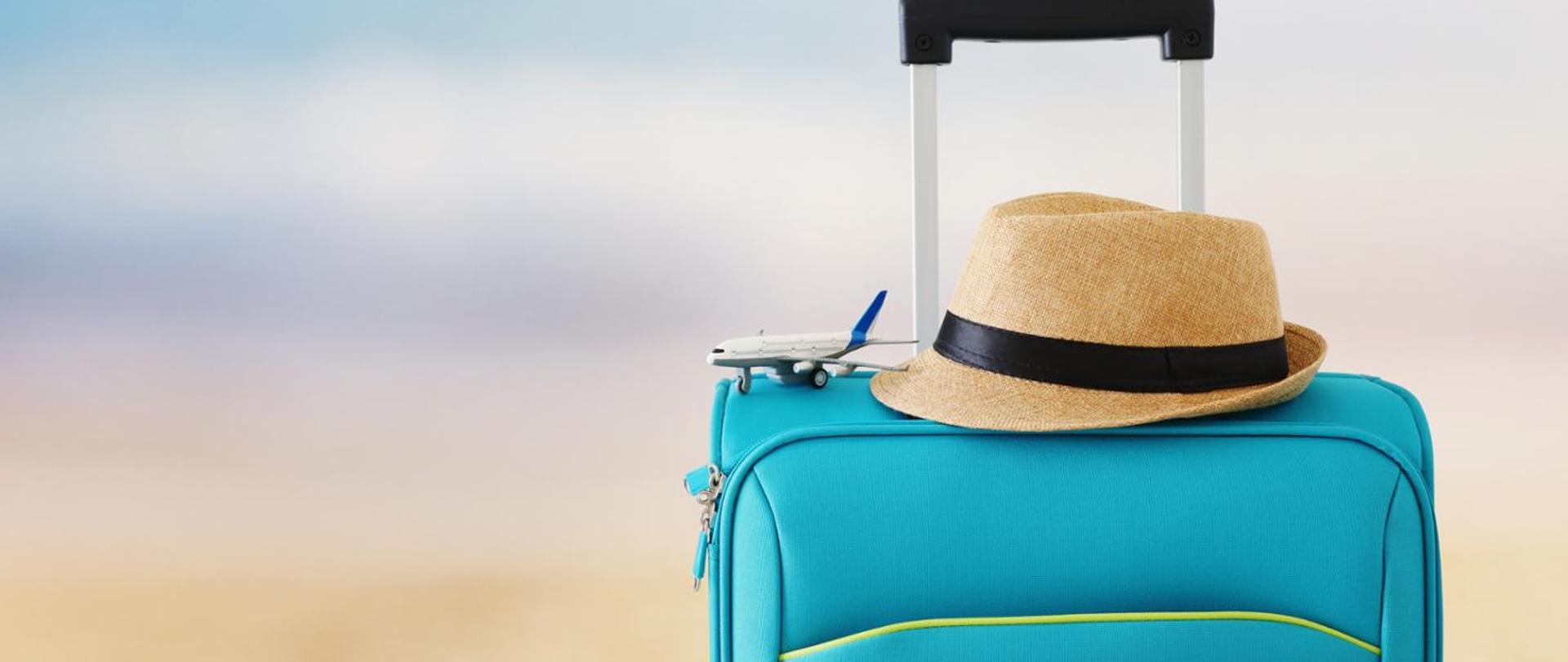 Błękitna walizka z czarną rączką. Na walizce żółty, letni kapelusz z czarną przepaską. Koło kapelusza mały samolot podróżny.