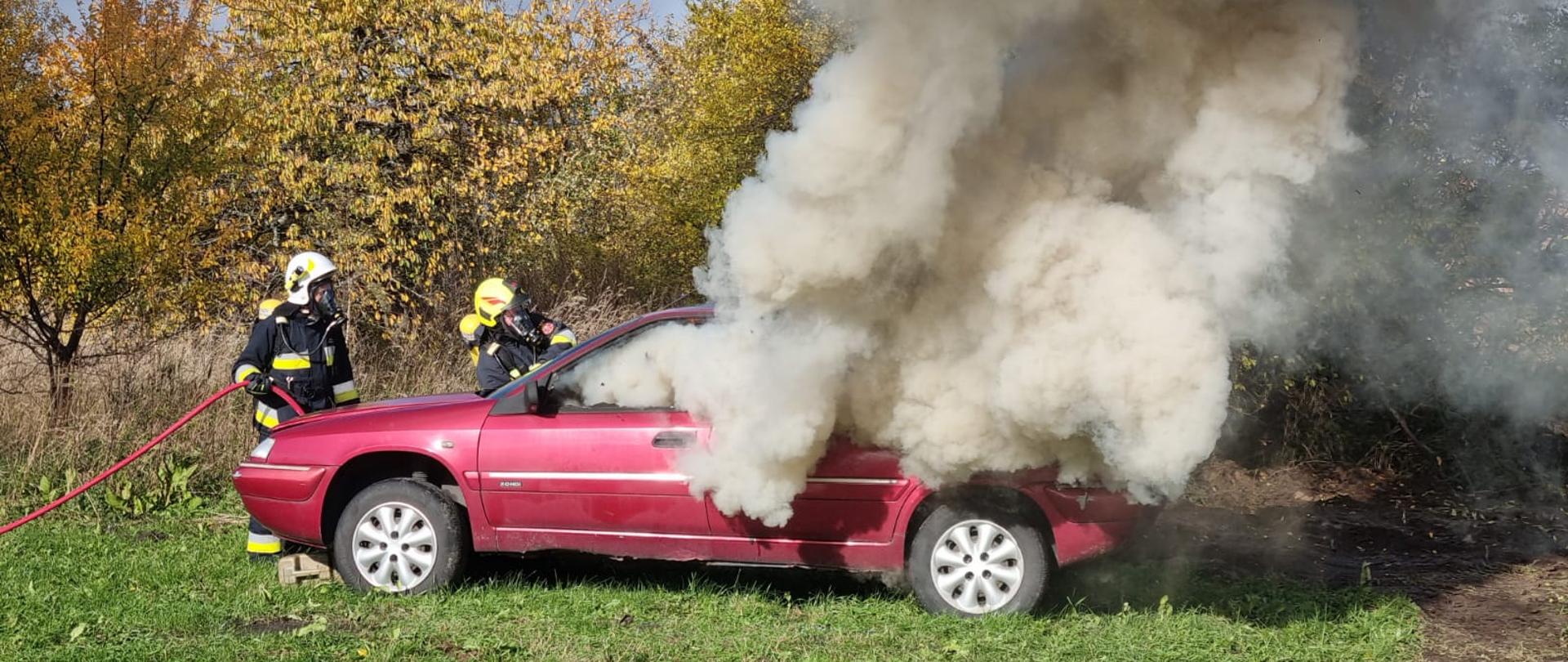 Zdjęcie przedstawia palący się czerwony samochód osobowy oraz dwóch strażaków gaszących go ubranych w ubrania specjalne oraz sprzęt ochrony układu oddechowego.