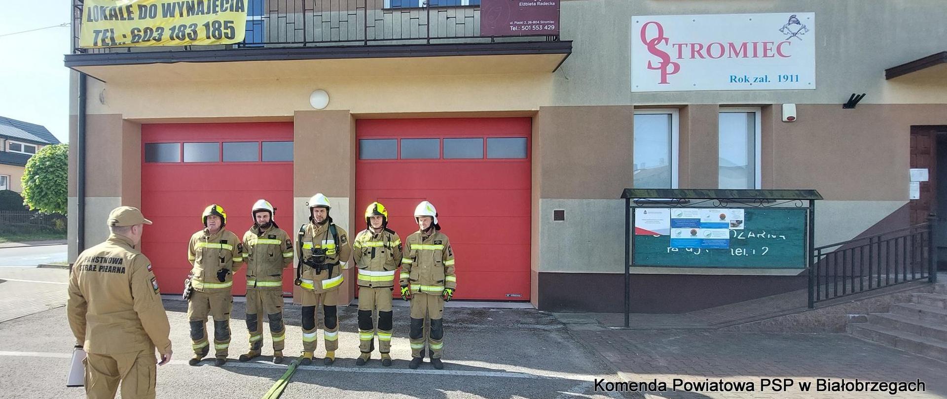 5 strażaków w piaskowych ubraniach specjalnych przed bramami garażowymi ochotniczej straży pożarnej w stromcu, przed nimi funkcjonariusz państwowej straży pożarnej w służbowym mundurze w kolorze piaskowym.