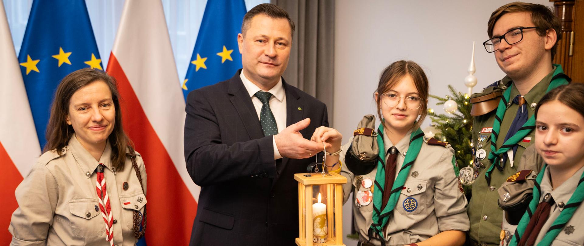 Pięć osób stoi, w tle flagi Polski i UE. Jedna z harcerek przekazuje ministrowi Paszykowi latarankę ze światełkiem 