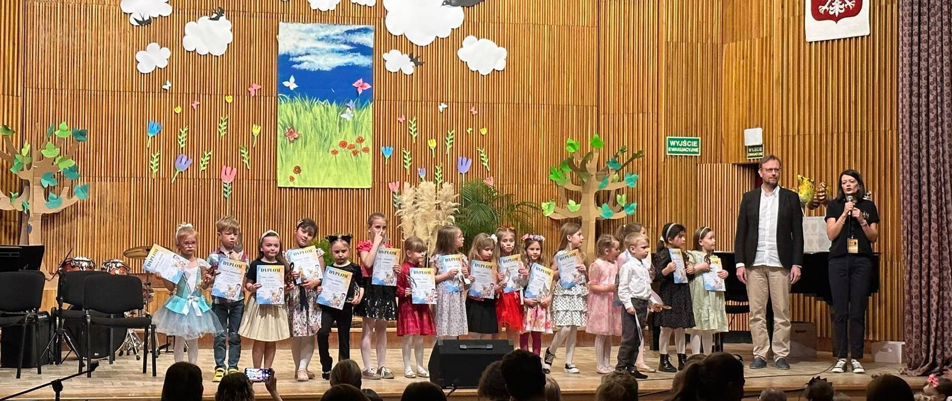 Widok na scenę sali koncertowej ZSM w Krośnie, na scenie kilkanaścioro dzieci oraz dwoje dorosłych. Dzieci trzymają w rękach dyplomy i statuetki, które otrzymały na rozdaniu nagród.