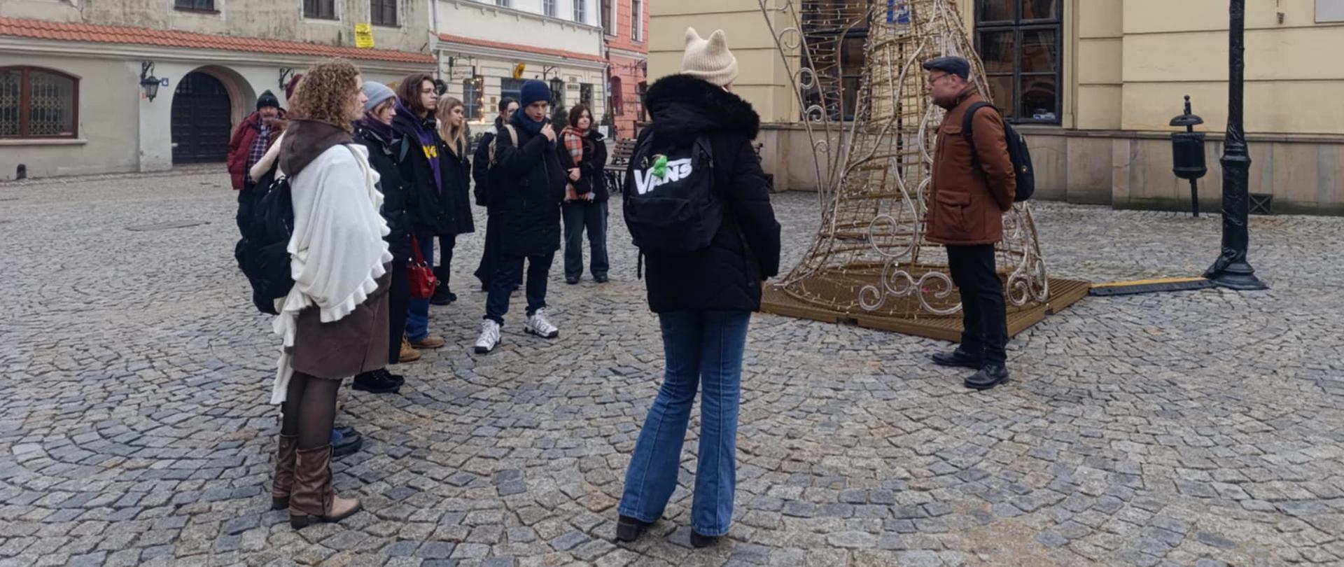 Jest bezsłoneczny, styczniowy dzień. Grupa uczniów i ich przewodnik Huber Mącik (mężczyzna w średnim wieku) stoją na Starym Mieście przed Trybunałem Koronnym, koło rzeźby - instalacji dużego anioła grającego na trąbce. Jest to dekoracja świąteczna (Boże Narodzenie). Uczniowie słuchają przewodnika.