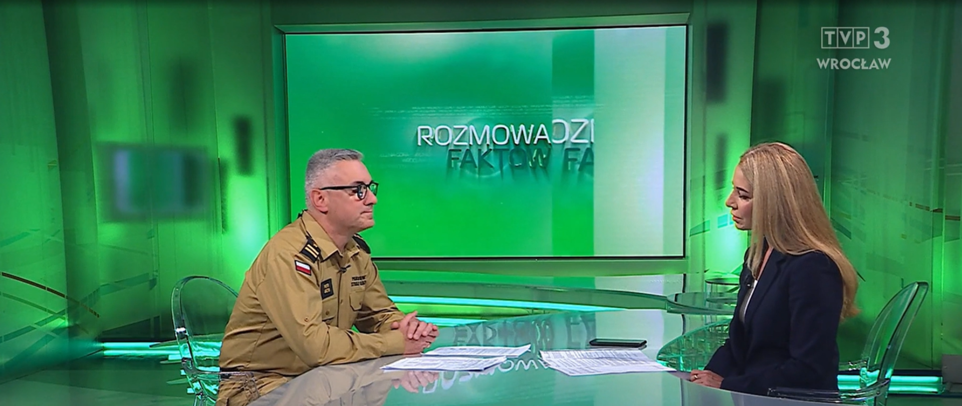 z-ca Komendanta Wojewódzkiego PSP we Wrocławiu siedzi przy stole i udziela wywiadu w telewizji. Obok siedzi osoba prowadząca program.