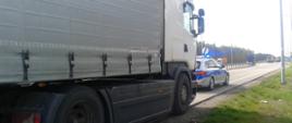 Na pierwszym planie (od lewej): ciągnik siodłowy z podpiętą naczepą typu firanka i oznakowany radiowóz Policji stojący przed zestawem ciężarowym. W tle: pojazdy poruszające się po ekspresowej „ósemce” w pobliżu Ostrowi Mazowieckiej.