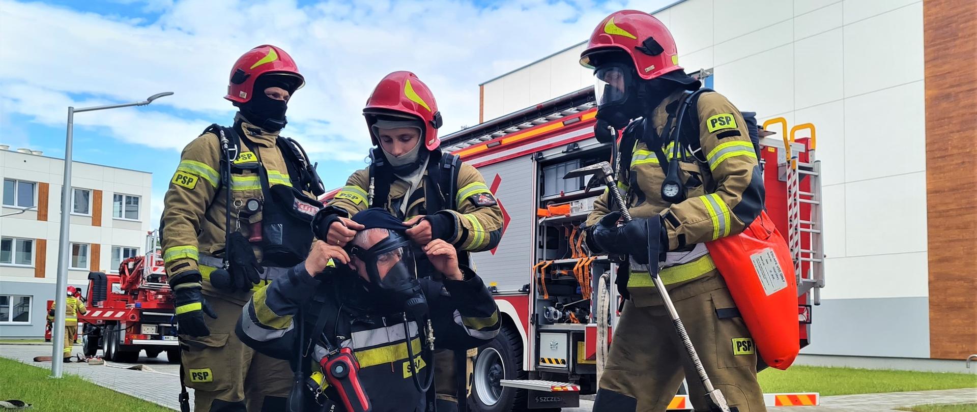 Zdjęcie przestawia strażaków podczas ćwiczeń. Strażacy ubrani są w stroje bojowe, na głowach mają czerwone hełmy. Na zdjęciu widoczne są również pojazdy pożanicze. Ćwiczenia odbywają się na obiekcie Szpitala Matki Bożej Nieustającej Pomocy w Wołominie