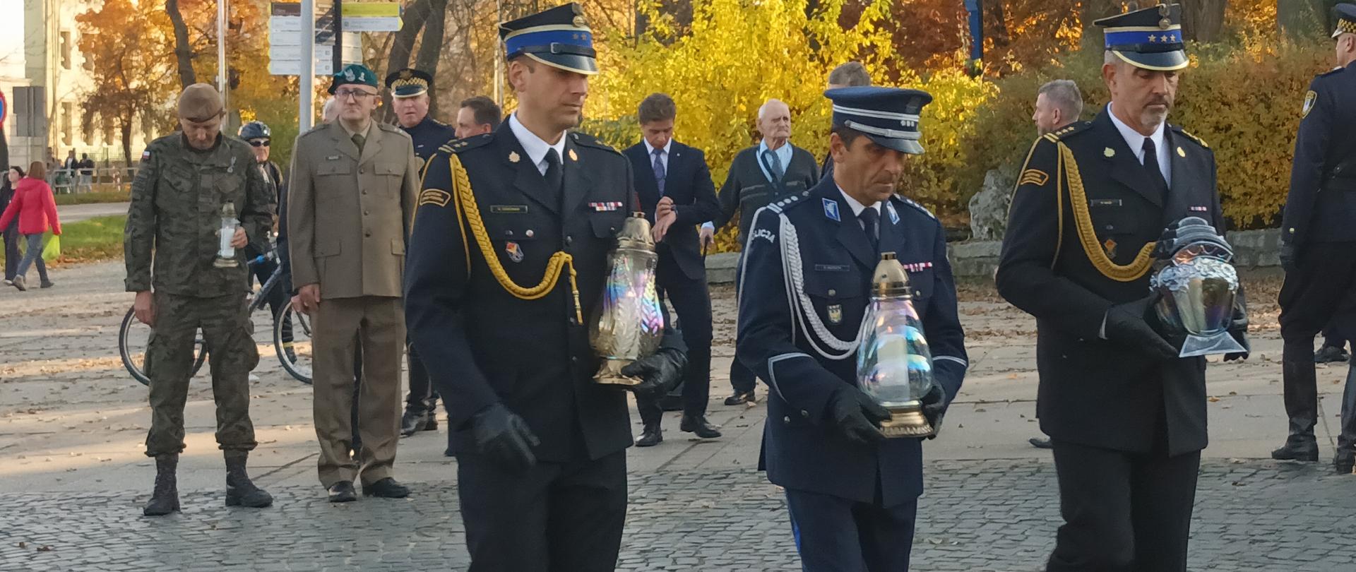 Oficer PSP, oficer Policji oraz Zastępca Komendanta Centralnej Szkoły PSP niosąc znicze podchodzą do grobu