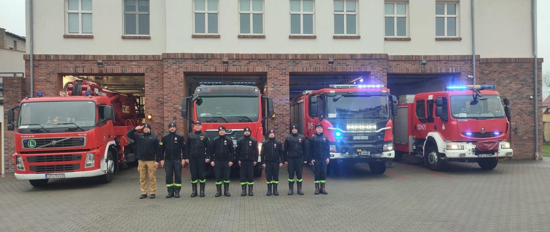 Strażacy ustawieni w szeregu oddają hołd ś.p. Piotrowi Mierneckiemu. Strażacy są ustawieni w szeregu ubrani w stroje koszarowe. Dowódca pierwszy z lewej w kolorze piaskowym reszta zaś w kolorze czarnym. Na drugim planie ustawione są wozy pożarnicze z JRG w Pucku.