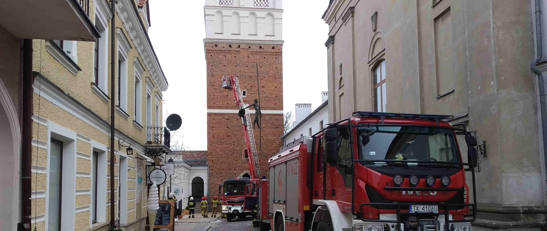 Zdjęcie zrobione w dzień. Na zdjęciu widać strażaków i samochody strażackie oraz sprawiony podnośnik do obiektu bramy opatowskiej. 