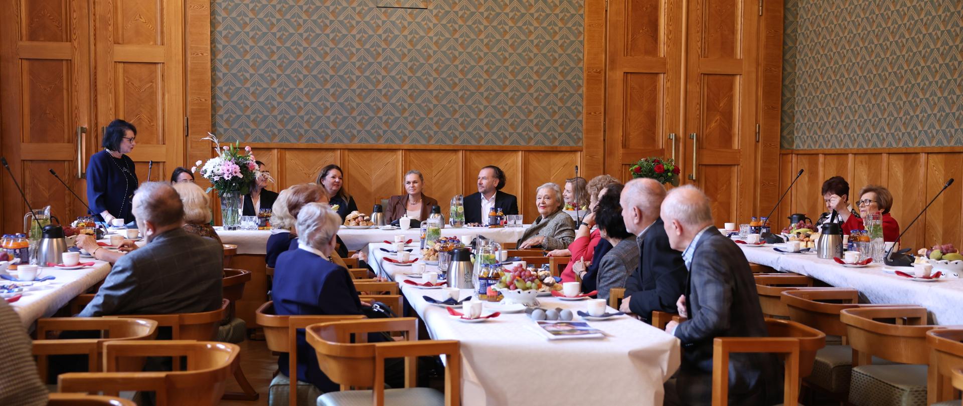 Duża sala ze ścianami w zielony wzorek, przy drewnianych stołach ustawionych w czworokąt siedzi dużo elegancko ubranych osób.