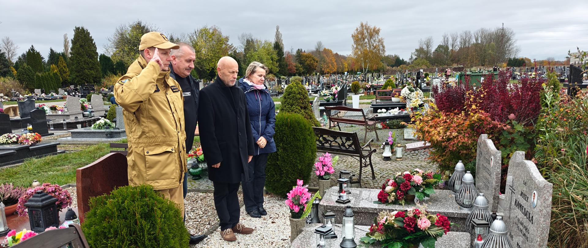 Strażak salutuje, inne osoby stoją w pozycji wyprostowanej nad grobem byłego strażaka