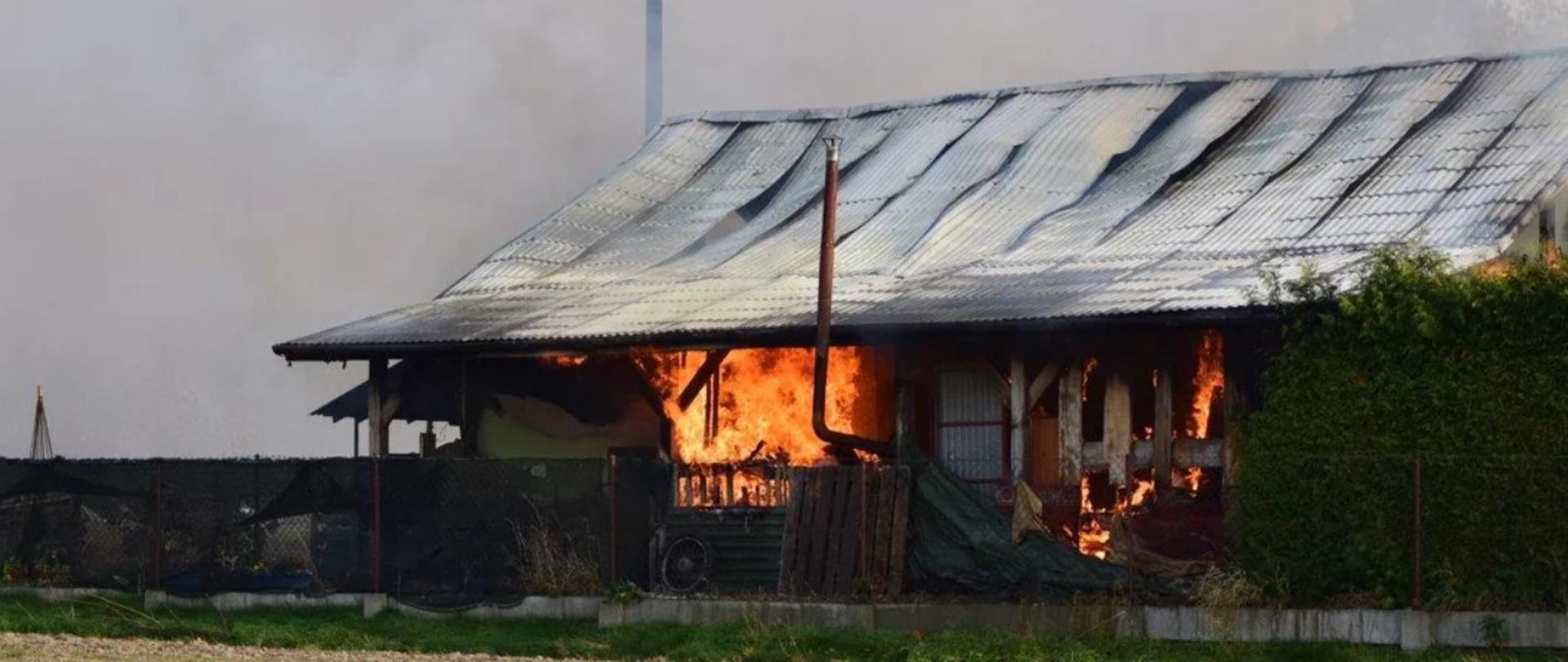 Zdjęcie palącego się budynku – w otworach drzwiowych i okiennych widać płomienie, na dachu blacha zdeformowana w wyniku działania wysokiej temperatury. Z całego poddasza wydobywa się dym.