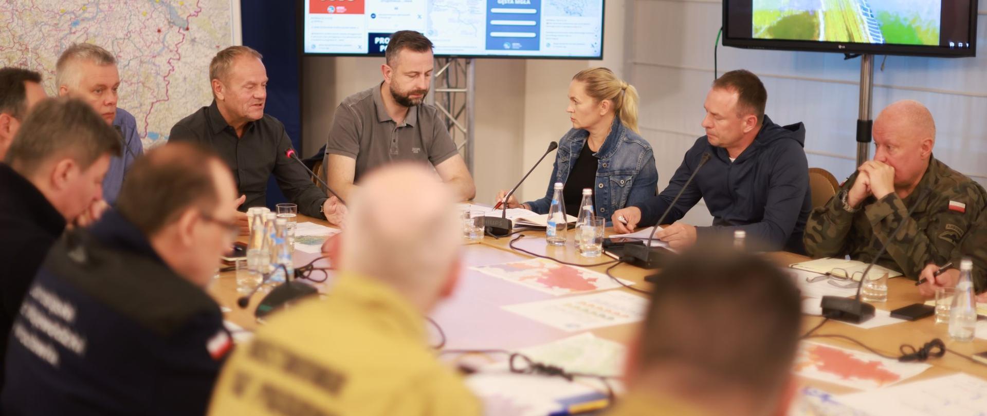 Ministra Edukacji uczestniczy w sztabie kryzysowym we Wrocławiu. Przy stole widać m.in. siedzącego premiera, ministra obrony narodowej, ministra spraw wewnętrznych i administracyjnych oraz innych członków władzy i służb. W tle widać zawieszoną mapę oraz dwa stojące ekrany z bieżącymi informacjami.