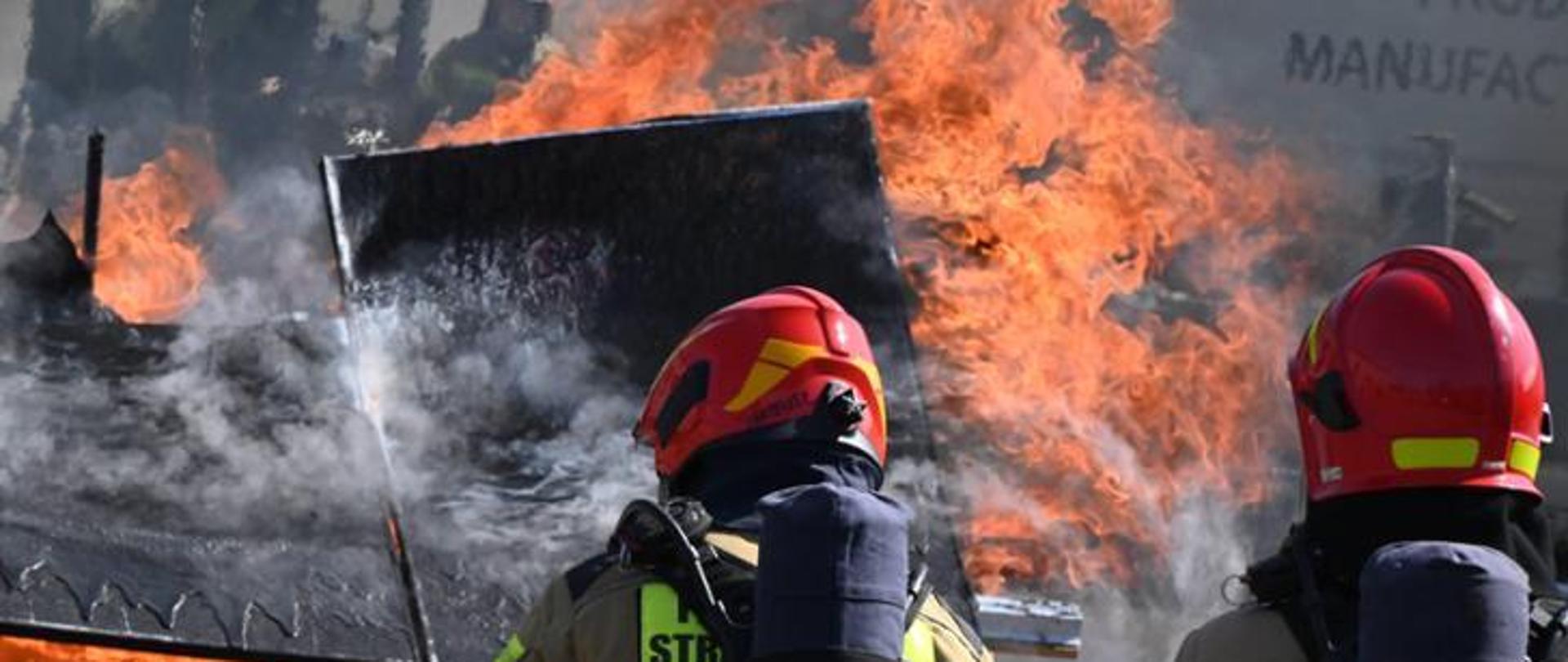 Dwaj strażacy odwróceni tyłem gaszą pożar panela fotowoltaicznego. Widoczne płomienie, dym i spalone materiały.
