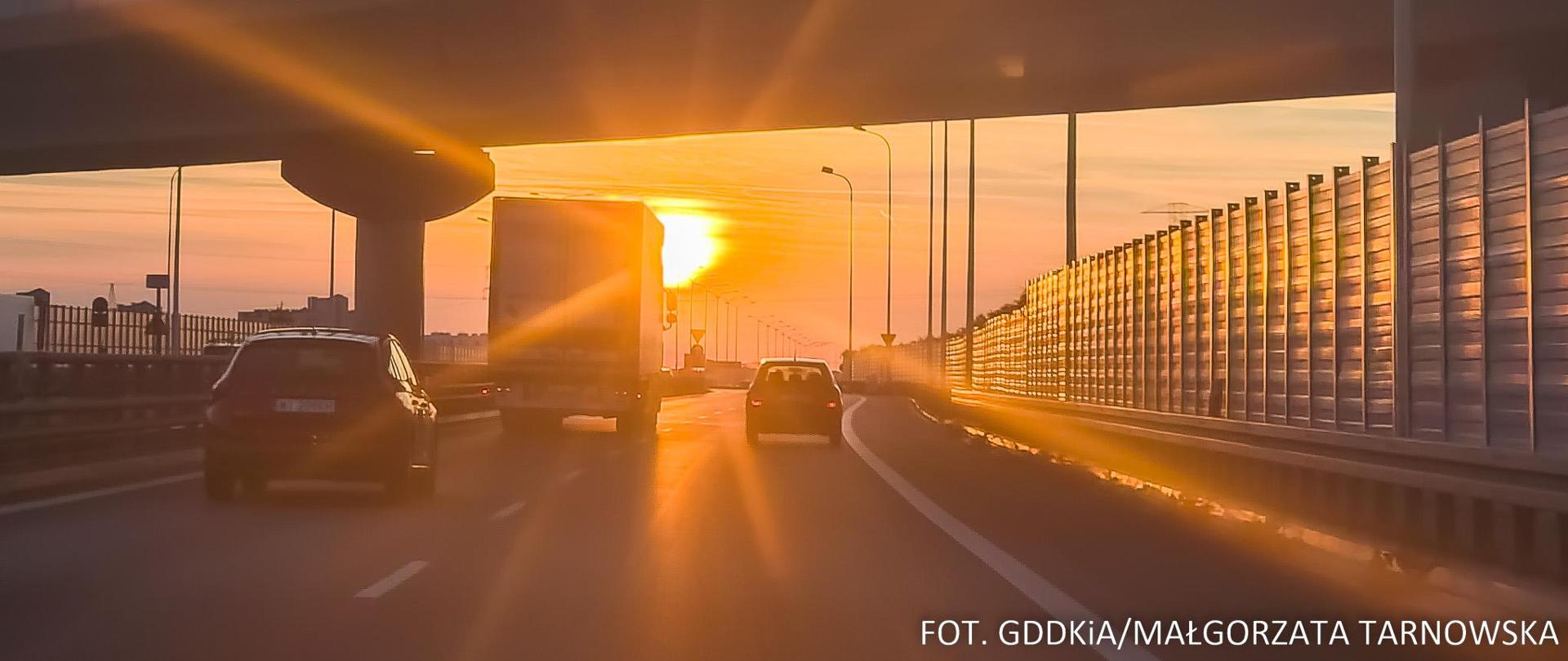 Widok z przodu samochodu na jezdnię drogi ekspresowej, po której jadą samochody. Na wprost świecące, zachodzące słońce.